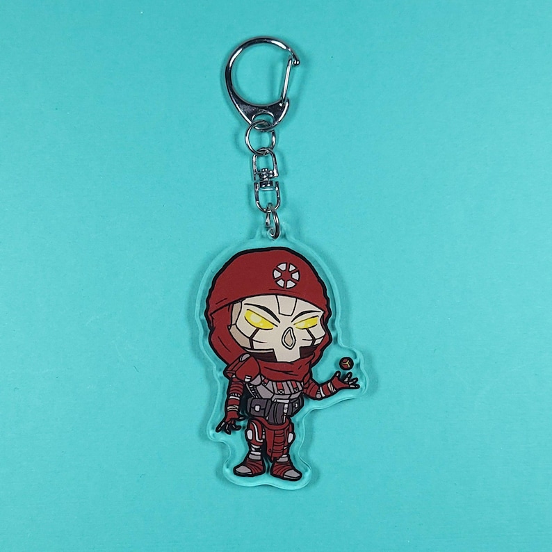 May include: A clear acrylic keychain featuring a cartoon character in a red hooded outfit with a skull-like face and yellow eyes. The keychain has a silver metal ring and chain. The character is holding a small black sphere.