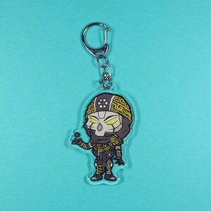 May include: A clear acrylic keychain featuring a cartoon character with a yellow and black design. The character has a helmet, mask, and is holding a small ball. The keychain has a silver metal clasp.
