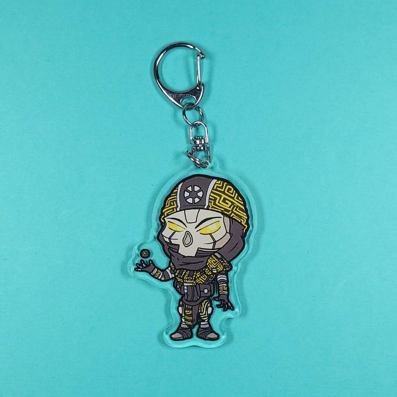 May include: A clear acrylic keychain featuring a cartoon character with a yellow and black design. The character has a helmet, mask, and is holding a small ball. The keychain has a silver metal clasp.