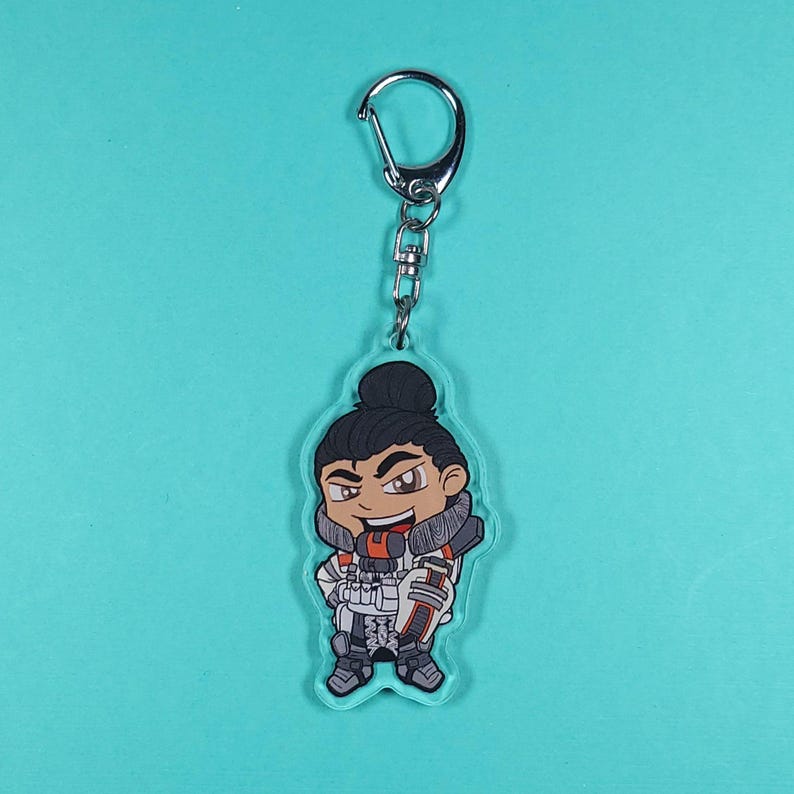 May include: A clear acrylic keychain featuring a cartoon character with brown hair, a white and orange outfit, and a black and white gun. The character is smiling and has a determined expression.