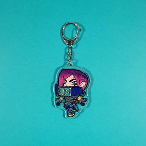 May include: A clear acrylic keychain featuring a cartoon character with pink hair, blue eyes, and a blue and gold costume. The character is wearing a mask and holding a weapon. The keychain is attached to a silver metal ring.