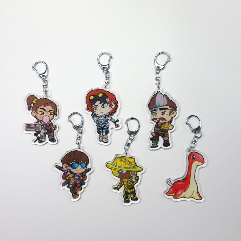 May include: Seven colourful cartoon character keychains with silver key rings. The characters include a woman with a gun, a man with a robot arm, a man with a beard, a man with a hat, a man with sunglasses, and a red dinosaur.