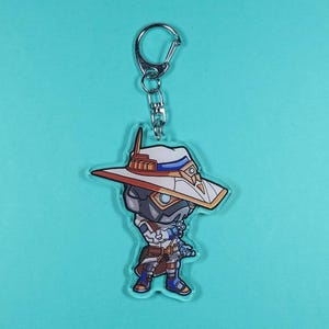 May include: A white and orange anime character keychain with a large hat and a silver key ring. The character is wearing a white and blue outfit and has a blue and white face.