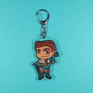 May include: A clear acrylic keychain featuring a cartoon character with brown hair, orange and red markings on her face, and a turquoise and brown outfit. She is holding a staff in her right hand and has a turquoise and brown belt.