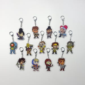 May include: A set of 13 colourful character keychains featuring characters from the video game Apex Legends. The keychains are made of clear acrylic and feature a variety of characters, including Bloodhound, Lifeline, and Mirage.