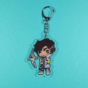 May include: A clear acrylic keychain featuring a cartoon character with brown hair, a white and black outfit, and a yellow and white weapon. The character is holding the weapon in their right hand and has a serious expression on their face.
