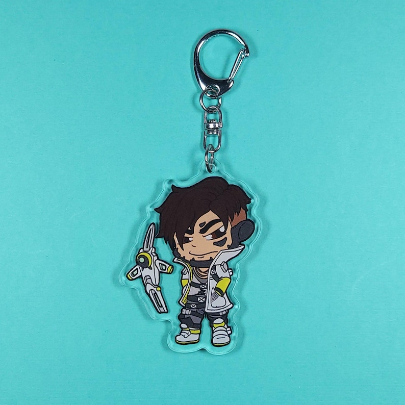 May include: A clear acrylic keychain featuring a cartoon character with brown hair, a white and black outfit, and a yellow and white weapon. The character is holding the weapon in their right hand and has a serious expression on their face.