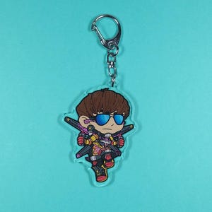 K&ouml;nnte beinhalten: Ein bunter Anime-Charakter-Schl&uuml;sselanh&auml;nger mit braunen Haaren, blauen Sonnenbrillen und einem rot-gelben Outfit. Der Charakter h&auml;lt eine Waffe und steht auf einem transparenten Acryl-Hintergrund.