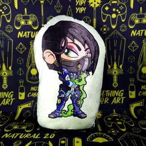 May include: A green plush pillow with a cartoon character of a woman with black hair, wearing a black and white mask, a blue and green outfit, and green and blue boots. The character is holding a green and blue weapon. The pillow has a white background and is surrounded by a black and yellow pattern.
