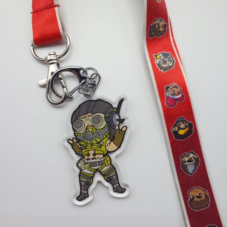 May include: A yellow and black character with a gas mask and goggles, wearing a yellow and brown outfit, is on a clear acrylic keychain. The keychain is attached to a silver key ring. A red and white lanyard with cartoon characters is in the background.