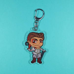 May include: A clear acrylic keychain featuring a cartoon illustration of a female character with red hair, wearing a white outfit and holding a weapon. The character has brown eyes and a determined expression.