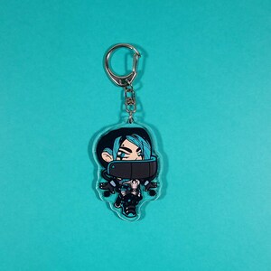 Op de afbeelding: Een transparante acryl sleutelhanger met een cartoonfiguur met blauw haar, een zwart masker en een blauw en zwart outfit. Het personage houdt een pistool in zijn rechterhand.