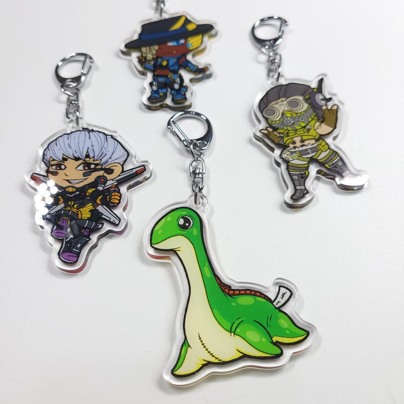 May include: Four different keychains featuring cartoon characters and a green dinosaur. The characters are a person with white hair and a yellow and black outfit, a person with a yellow and black outfit and a hat, a person with a yellow and green outfit and goggles, and a green dinosaur with a red stripe down its back.