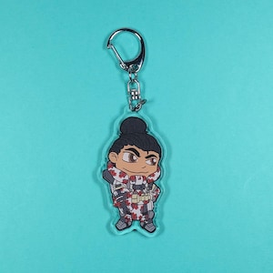 May include: A clear acrylic keychain featuring a chibi-style illustration of a video game character. The character is wearing a white and red camouflage outfit with a black and white bandana. The character has black hair in a bun and brown eyes.