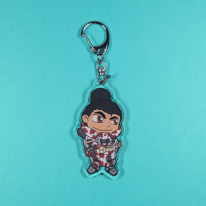 May include: A clear acrylic keychain featuring a chibi-style illustration of a video game character. The character is wearing a white and red camouflage outfit with a black and white bandana. The character has black hair in a bun and brown eyes.