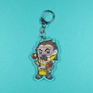 May include: A clear acrylic keychain featuring a cartoon illustration of a character from the video game Apex Legends. The character is wearing a yellow and black gas mask and a yellow and brown jumpsuit. The character is holding a red and white can in their left hand.