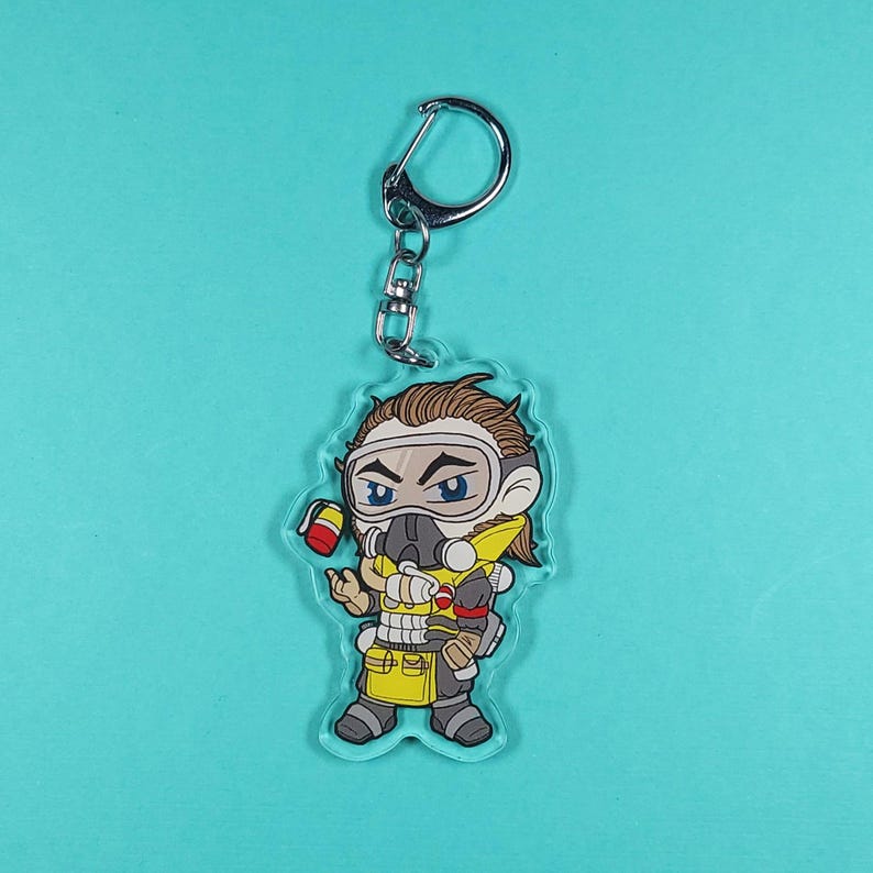 May include: A clear acrylic keychain featuring a cartoon illustration of a character from the video game Apex Legends. The character is wearing a yellow and black gas mask and a yellow and brown jumpsuit. The character is holding a red and white can in their left hand.