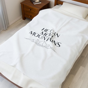 May include: White blanket featuring the text "HE CAN MOVE MOUNTAINS" in black, accompanied by a mountain illustration. The blanket is on a bed with a white pillow. A wooden bedside table is visible.
