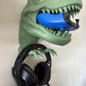 May include: A green dinosaur head wall mount holding a blue game controller in its mouth and a black headset on its claws. The dinosaur has yellow eyes and sharp teeth. A fun and functional gaming accessory.