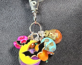 May include: A Halloween-themed keychain with a silver clasp. The charm features a yellow crescent moon with a black bat, purple glitter, and orange and teal skull beads. Pink and orange beads complete the design.
