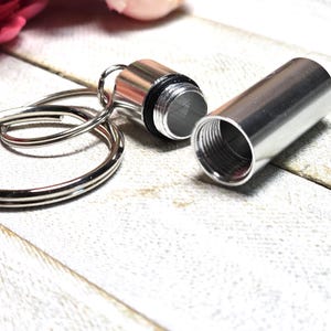 May include: A silver metal keychain pill container with a keyring. The container is open and the lid is off, revealing the inside of the container.