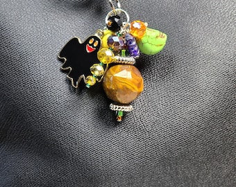 May include: A Halloween-themed keychain with a black ghost charm, a large, round, marbled bead, and colorful beads. The keychain is attached to a black bag with a silver zipper.