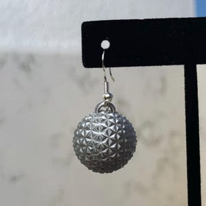 May include: A single silver-colored earring with a textured, spherical design. The earring hangs from a silver hook and is displayed against a black background. The sphere has a geometric pattern.