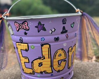 May include: Purple Halloween trick-or-treat bucket with the name "Eden" written in yellow with orange stripes. The bucket is decorated with stars, a ghost, a witch's hat, and other Halloween-themed designs.