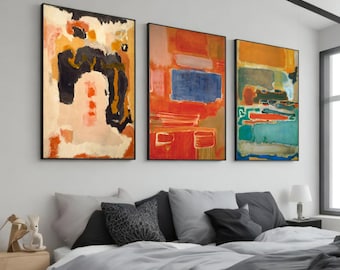 Op de afbeelding: Drie abstracte schilderijen in zwarte lijsten, met een mix van rode, oranje, gele, blauwe en groene kleuren. De schilderijen hangen aan een witte muur boven een grijs bed met zwarte en grijze kussens.