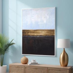 Puede incluir: Pintura abstracta de paisaje con un cielo azul, un suelo texturizado dorado y marrón y un fondo marrón oscuro. La pintura está enmarcada en un marco blanco.