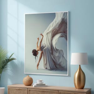 Op de afbeelding: Een ingelijste kunstdruk met een ballerina in een vloeiende witte jurk, vastgelegd in beweging. De danseres staat op spitzen, met sierlijk uitgestrekte armen. De achtergrond is een zachte gradiënt van blauw en wit, wat een serene sfeer creëert.