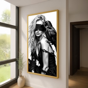 May include: A framed black and white artwork depicts a woman with long blonde hair and a blindfold, with a black raven perched on her shoulder. The artwork is in a gold frame and hangs on a white wall.