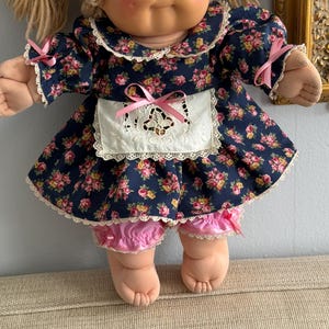May include: A vintage doll with blonde yarn hair tied with pink ribbons. The doll wears a navy blue floral dress with a white apron and pink bloomers. The dress has a white lace trim.