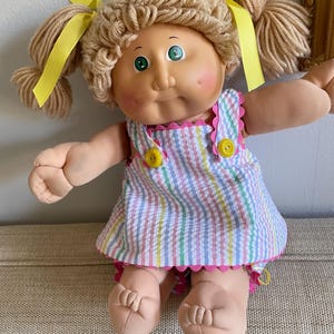 May include: A vintage Cabbage Patch Kids doll with blonde yarn hair styled in pigtails, adorned with yellow ribbons. The doll wears a seersucker dress with pink and yellow accents and yellow buttons.