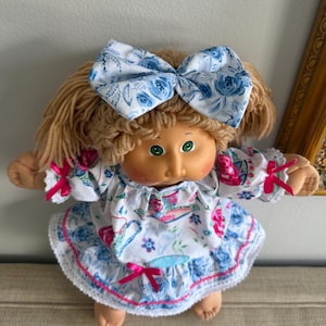 May include: A vintage doll with blonde yarn hair, green eyes, and a painted face. The doll wears a white dress with a blue floral pattern, pink accents, and a matching bow. The dress has a ruffled skirt and sleeves.