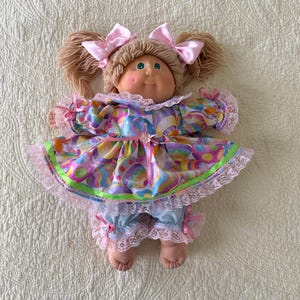May include: A vintage doll with a round face, green eyes, and light brown yarn hair styled in pigtails with pink bows. The doll wears a colorful dress with a pink and green trim, lace trim, and blue bloomers with lace trim.