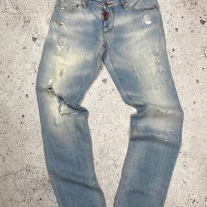 May include: Light blue distressed denim jeans with faded areas and several rips and tears. The jeans have a button fly and a red tag on the back pocket.