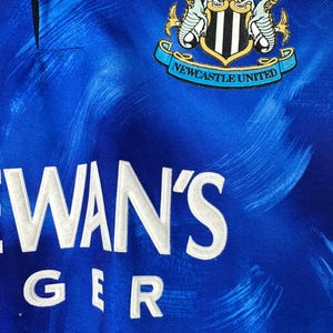 May include: A blue football shirt with a white and black crest that says "Newcastle United". The shirt has a white sponsor logo that says "NEWAN'S GER".