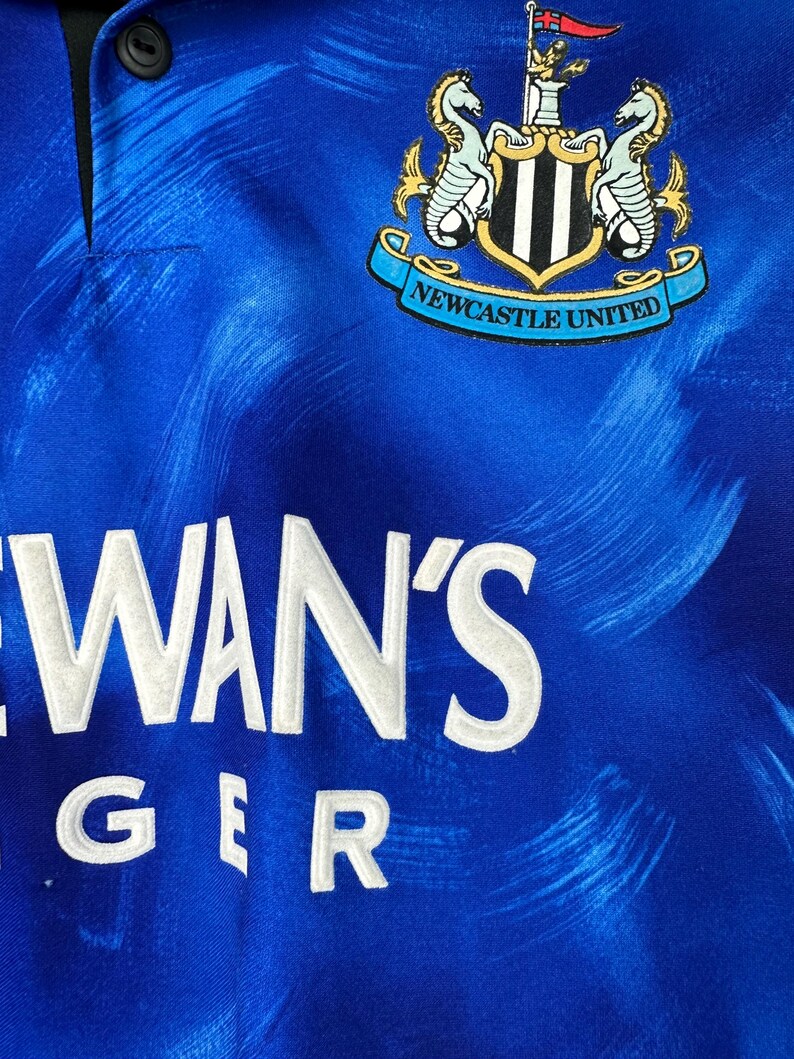 May include: A blue football shirt with a white and black crest that says "Newcastle United". The shirt has a white sponsor logo that says "NEWAN'S GER".