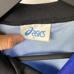 May include: A blue and black athletic shirt with the Asics logo and the size XL on a white tag.