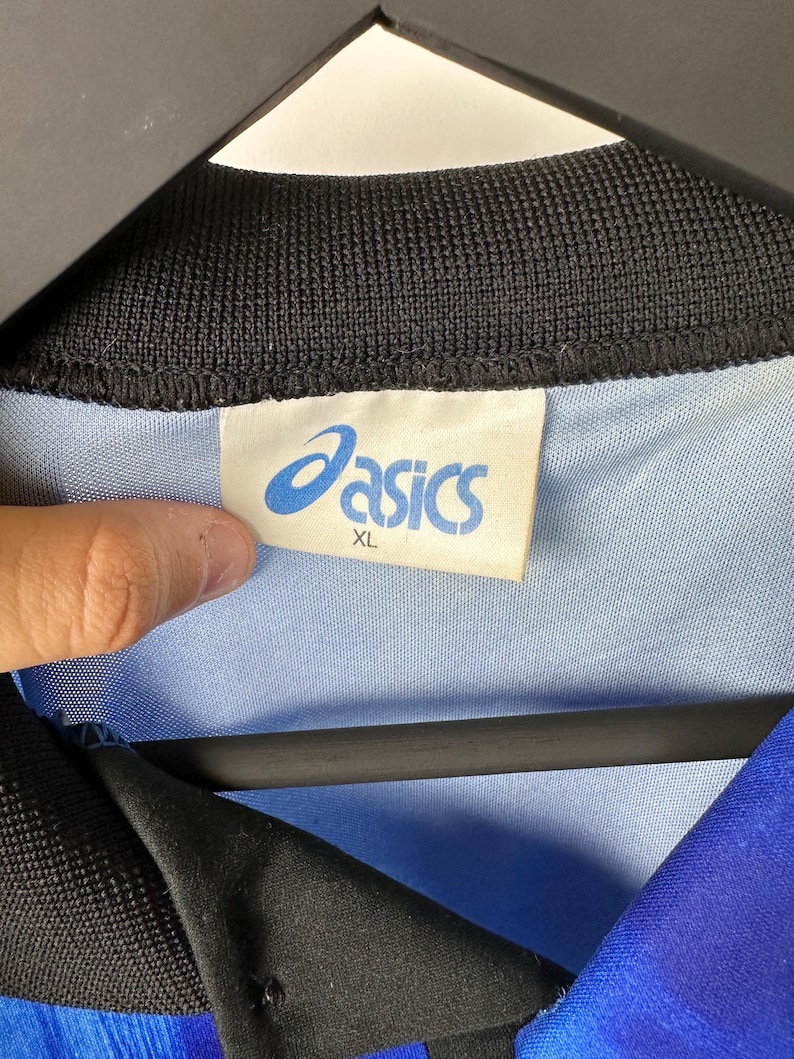 May include: A blue and black athletic shirt with the Asics logo and the size XL on a white tag.