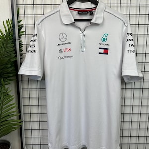 Może przedstawiać: Biała koszulka polo z krótkim rękawem i zamkiem błyskawicznym. Koszulka ma kilka logo, w tym Mercedes-AMG, UBS, Petronas i Qualcomm.