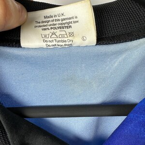May include: A white tag with black text that reads "Made in U.K. The design of this garment is protected under copyright law 100% POLYESTER" with care instructions below. The tag is attached to a blue and black shirt.