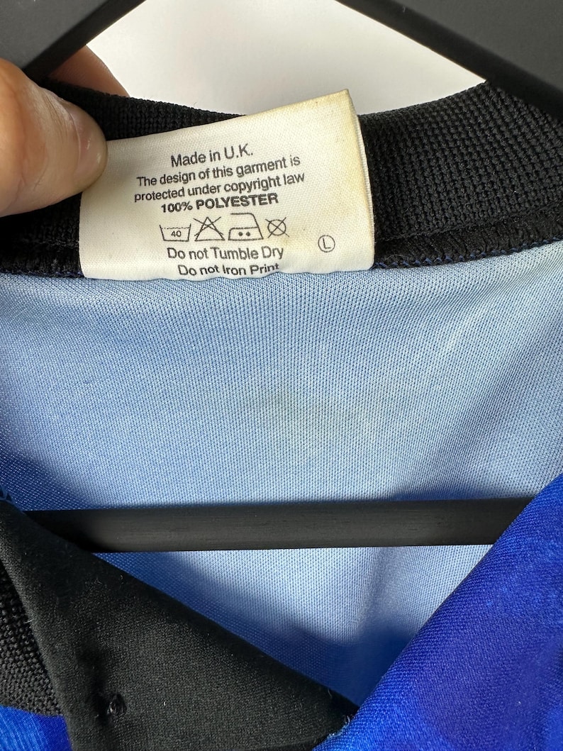 May include: A white tag with black text that reads "Made in U.K. The design of this garment is protected under copyright law 100% POLYESTER" with care instructions below. The tag is attached to a blue and black shirt.
