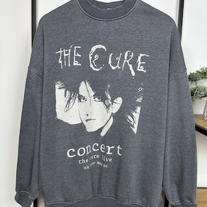 Könnte beinhalten: Ein anthrazitfarbenes Sweatshirt mit einem weißen Aufdruck der Band "The Cure" und dem Wort "Konzert". Die Grafik enthält ein Porträt und den Text "the cure live uk may 84". Das Sweatshirt hat einen Rundhalsausschnitt und lange Ärmel.
