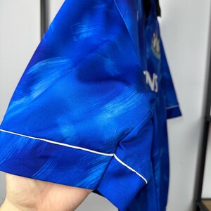 May include: A blue football shirt with a white trim and a faded blue and white pattern. The shirt has a white logo on the left chest and the text "NS" on the right side.
