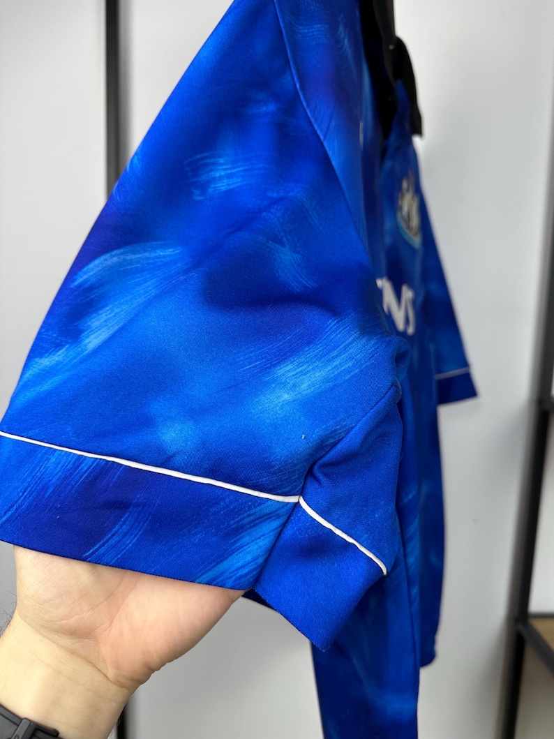 May include: A blue football shirt with a white trim and a faded blue and white pattern. The shirt has a white logo on the left chest and the text "NS" on the right side.