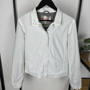 Op de afbeelding: Een witte jas met kraag en knoopsluiting. De jas heeft lange mouwen en twee zakken. Het label luidt "Art. 108173 Mat. 62T". De jas hangt aan een zwarte hanger.