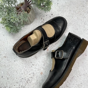 May include: A pair of black leather Mary Jane shoes with a buckle strap and a brown rubber sole.