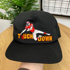 Peut inclure: Casquette de baseball noire avec une image brodée d'un joueur de football américain marquant un touché. Le texte "TOUCHDOWN" est brodé en orange et jaune sur le devant de la casquette.